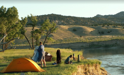 Two campers set up tents by a river, surrounded by green grass and hills under a clear sky.