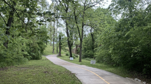 A winding path through a lush green park, surrounded by trees and grass, with signs along the trail.