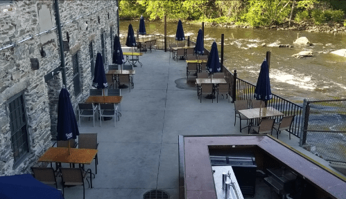 Outdoor dining area by a river, featuring tables, chairs, and umbrellas along a stone building.