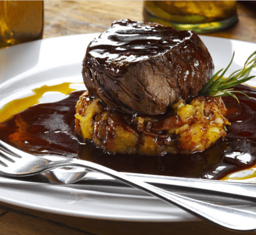 A gourmet dish featuring a tender beef filet on a bed of savory mashed potatoes, drizzled with rich sauce and garnished.