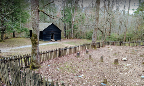 A rustic black cabin surrounded by trees, with a fenced graveyard featuring stone markers in the foreground.
