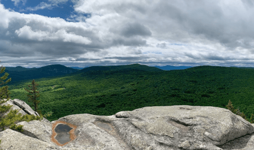 A panoramic view of lush green mountains under a cloudy sky, with rocky foreground and distant peaks.