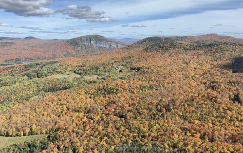 A panoramic view of vibrant autumn foliage covering rolling hills under a partly cloudy sky.