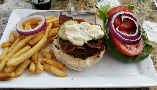 A plate with a burger topped with bacon, pickles, and mayonnaise, served with fries and fresh vegetables on the side.