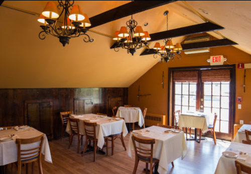 Cozy restaurant interior with wooden beams, chandeliers, and tables set for dining near a door leading outside.