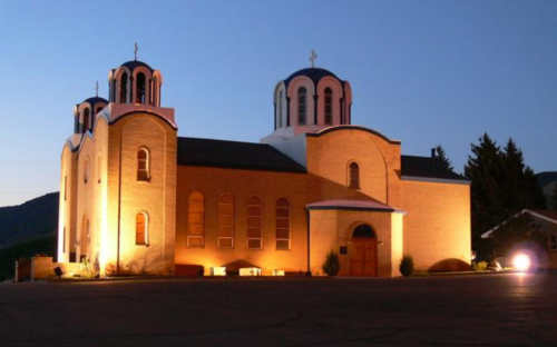 A lit-up church with two domes at dusk, surrounded by trees and mountains in the background.