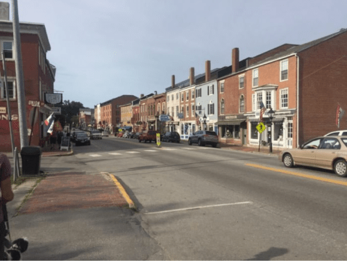 A street view of a small town with brick buildings, parked cars, and flags along the sidewalk.