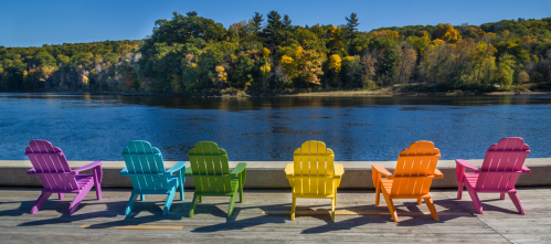 Colorful Adirondack chairs line a wooden deck by a river, surrounded by autumn foliage and a clear blue sky.
