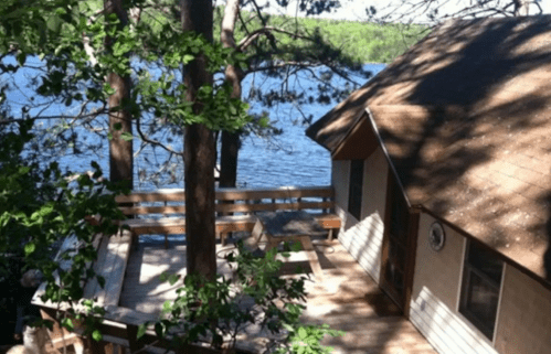 A cozy cabin by the lake, surrounded by trees and featuring a wooden deck overlooking the water.