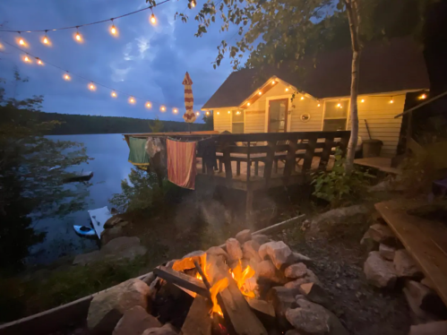 A cozy lakeside cabin at dusk, with string lights, a fire pit, and colorful towels hanging on a railing.