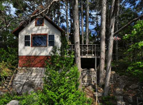 A quaint house nestled among tall trees, featuring a deck and stone steps, with an American flag visible nearby.