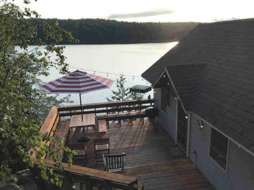 A lakeside deck with a striped umbrella, table, and chairs, overlooking calm water and trees at sunset.