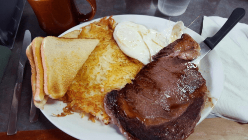 A hearty breakfast plate with a steak, eggs, hash browns, and toast, accompanied by a coffee mug and water glass.