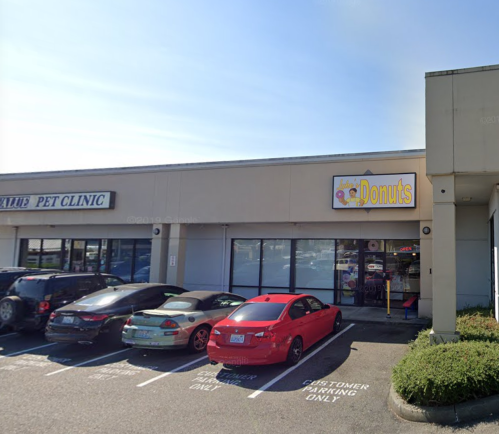 A storefront with a "Donuts" sign, next to a pet clinic, featuring parked cars in front. Bright, sunny day.
