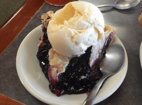 A slice of blueberry pie topped with a scoop of vanilla ice cream on a small white plate.