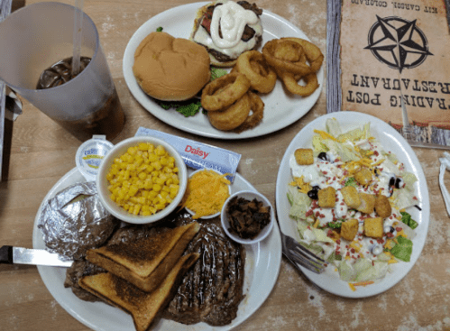 A table with a burger, onion rings, a salad, corn, cheese, and a steak with toast, accompanied by a drink.