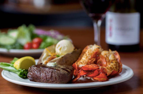 A plate featuring steak, lobster, baked potato, and salad, with a glass of red wine in the background.