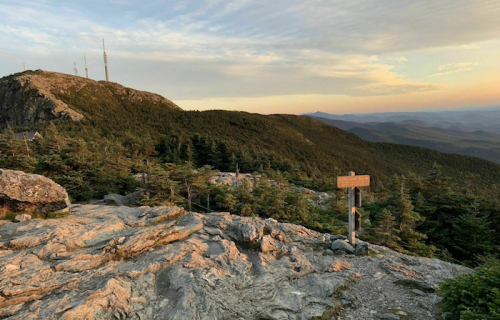 A rocky mountain landscape at sunset, with trees and a signpost in the foreground and distant mountains in the background.