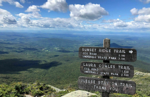 Signpost at a mountain trail with directions to Sunset Ridge and Laura Cowles Trails, overlooking a scenic landscape.