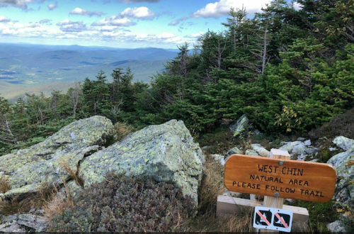 A wooden sign at a mountain peak reads "West Chin Natural Area, Please Follow Trail," with rocky terrain and trees in the background.