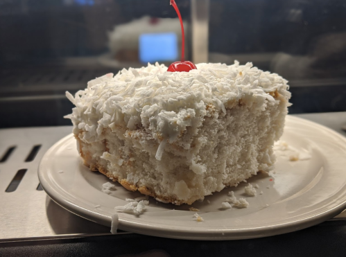 A slice of coconut cake topped with shredded coconut and a cherry, served on a white plate.