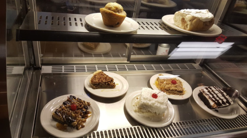 A display case with various desserts on white plates, including cakes, pies, and pastries, topped with cherries.
