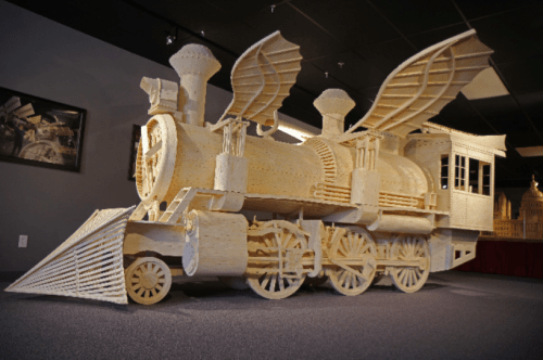 A large, intricately crafted wooden train model with wings, displayed in a gallery setting.
