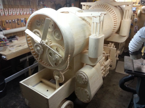 A large, intricate wooden model of a steam engine with visible gears and mechanical components in a workshop setting.