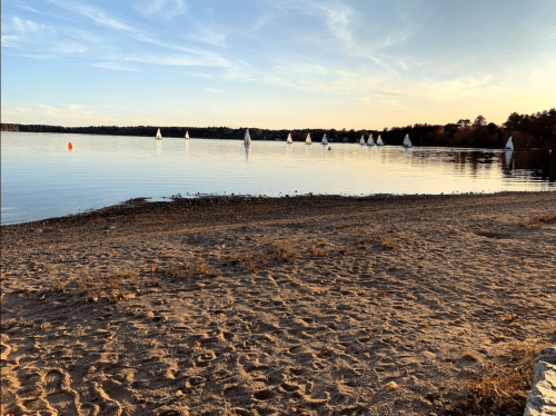 A serene lakeside view at sunset, with sailboats on calm water and a sandy shore in the foreground.