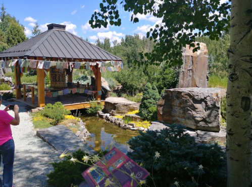 A serene garden scene featuring a gazebo with prayer flags, a pond, and a rock waterfall surrounded by greenery.