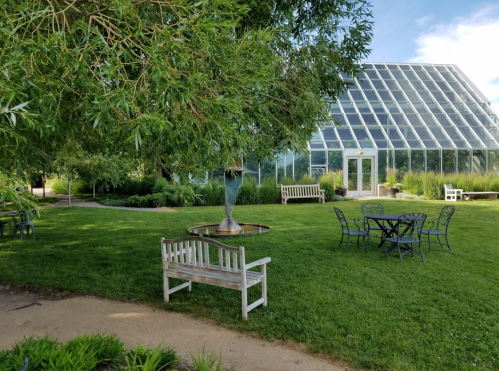 A serene garden with a greenhouse, a fountain, benches, and a table surrounded by lush greenery under a blue sky.