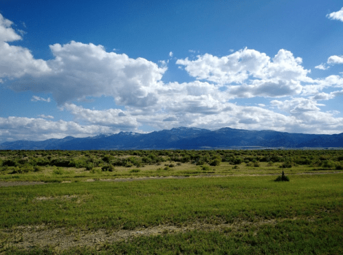 A wide landscape featuring green fields, distant mountains, and a blue sky with fluffy white clouds.