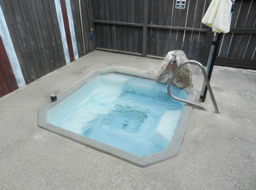 A small octagonal hot tub with clear water, surrounded by a concrete patio and wooden fence.