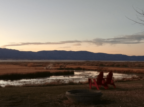 A serene landscape at sunset featuring mountains, a pond, and red Adirondack chairs in the foreground.