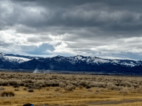 A dramatic landscape featuring snow-capped mountains under a cloudy sky, with a grassy foreground and distant smoke.