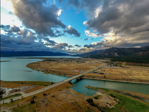 Aerial view of a bridge over a river, surrounded by fields and mountains under a cloudy sky.