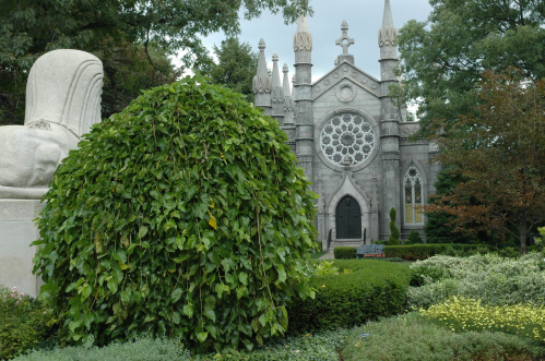A stone building with a rose window, surrounded by lush greenery and a sculpted figure in a garden setting.