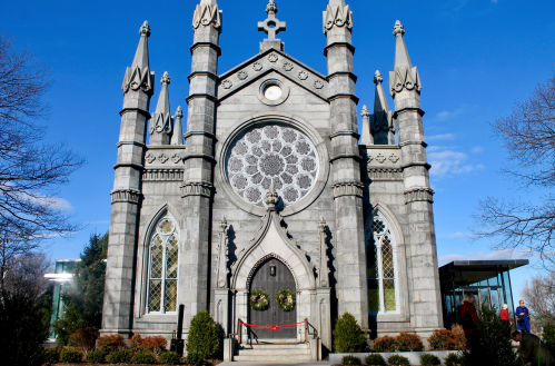 A gothic-style stone chapel with a large rose window, surrounded by greenery and a clear blue sky.