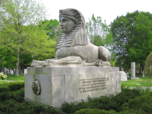 A large stone sphinx statue in a cemetery, surrounded by greenery and gravestones.