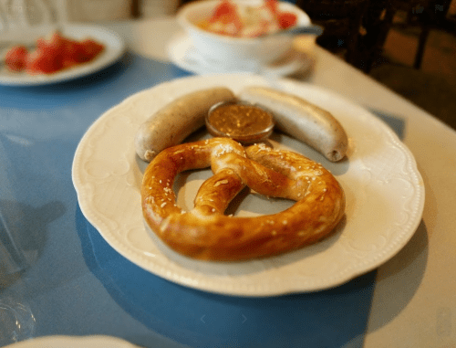 A plate with a large pretzel, two sausages, and a small bowl of mustard.
