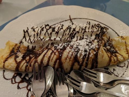 A delicious crepe drizzled with chocolate sauce and dusted with powdered sugar, surrounded by small spoons.