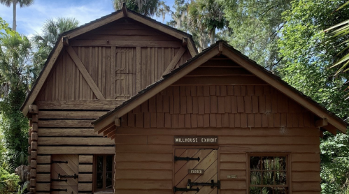 A rustic wooden building surrounded by greenery, featuring a sign that reads "Millhouse Exhibit" and marked "Closed."