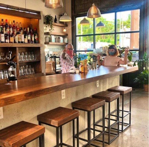 Two people sit at a bar with a wooden counter, surrounded by plants and a well-stocked shelf of drinks.