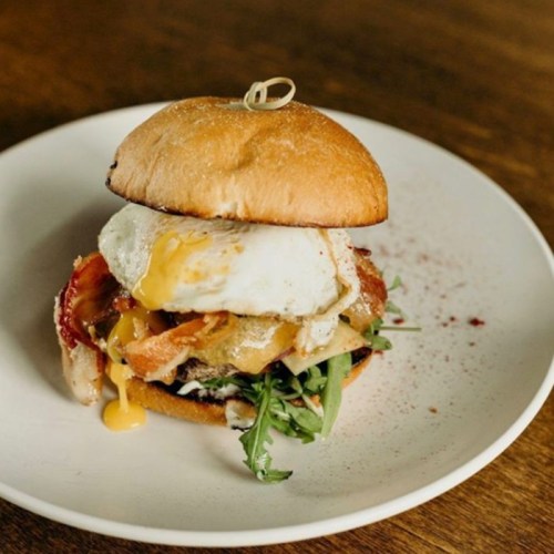 A delicious burger with a sunny-side-up egg, bacon, greens, and a toasted bun on a white plate.