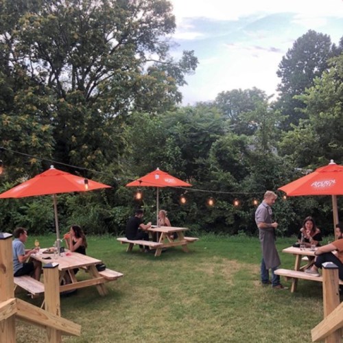 Outdoor dining area with wooden picnic tables, orange umbrellas, and string lights, featuring people enjoying meals.