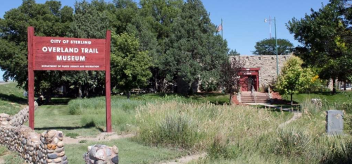 Sign for the Overland Trail Museum in Sterling, surrounded by greenery and a stone wall.