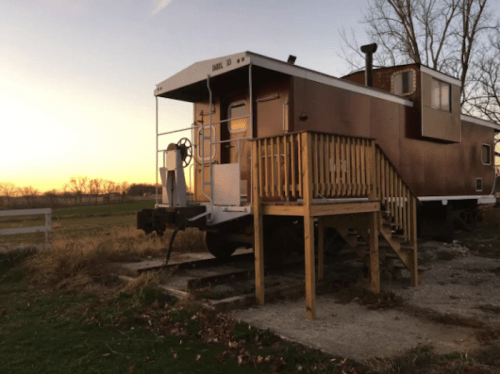 A vintage train caboose converted into a cozy cabin, set against a sunset backdrop and surrounded by grassy fields.