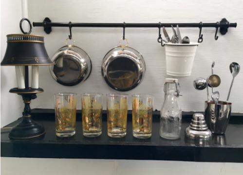 A shelf displaying a lamp, pots, utensils, and five glasses with a golden design, alongside a glass bottle and a shaker.
