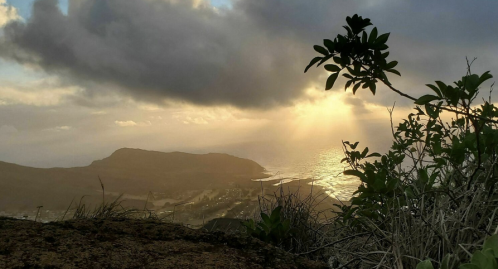A scenic view of the ocean at sunset, with clouds and a silhouette of a mountain in the background.