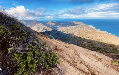 A person sits on a rocky hillside, overlooking a lush valley and the ocean under a blue sky with scattered clouds.